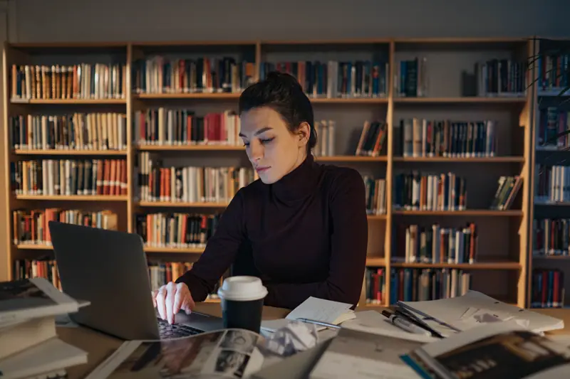Voksen kvinne som arbeider konsentrert med bærbar PC i et bibliotek med bokhyller